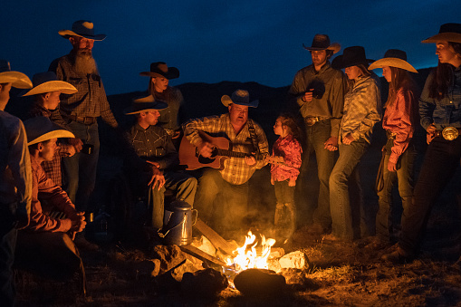 Large Group of cowboys and cowgirls together after a long outdoor day relaxing at the campfire together. Cowboy singing and playing guitar. Utah, USA.
