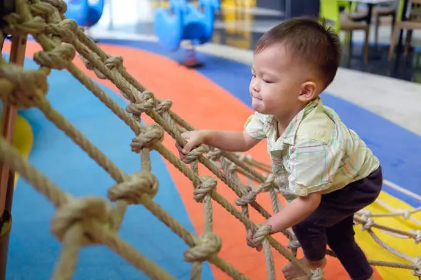 Photo of Cute little Asian 2 years old toddler baby boy child having fun trying to climb on jungle gym at indoor playground