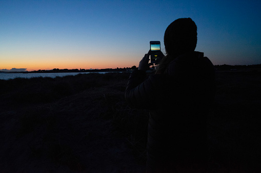 A woman taking a picture of the sunset with her mobile phone.