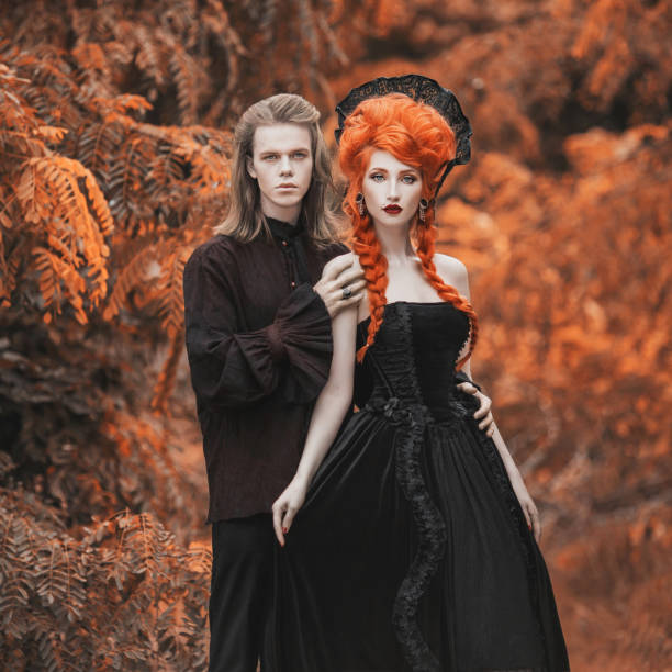 Gothic couple in halloween costume. Gloomy vampire in victorian clothes. Redhead woman vampire in black victorian dress. Gothic clothes for halloween party. Gloomy couple on autumn background. Gothic couple in halloween costume. Gloomy vampire in victorian clothes. Redhead woman vampire in black victorian dress. Gothic clothes for halloween party. Gloomy couple on autumn background. gothic fashion stock pictures, royalty-free photos & images