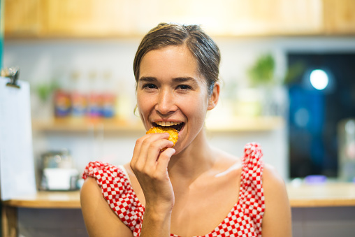 Portrait of happy young woman eating cookie in kitchen. Latin American woman biting pastry with pleasure. Food concept