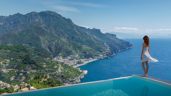 Italy. Amalfi Coast. A young woman standing on the edge of  an infinity pool of a private luxury villa of Ravello watching the coastline .