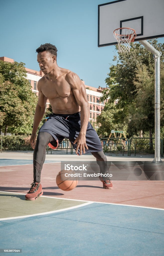Afroamerican young man playing street basketball in the park One afroamerican young man without tshirt is playing basketball in a park in Madrid during summer at midday. He is bouncing the ball under the basket. Active Lifestyle Stock Photo