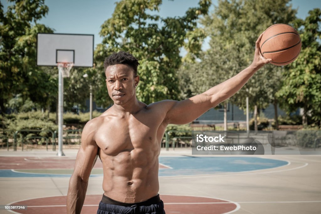 Afroamerican young man playing street basketball in the park Close up of one afroamerican young man without tshirt playing basketball in a park in Madrid during summer at midday. He is standing and holding the ball with one hand loooking at the camera. Active Lifestyle Stock Photo