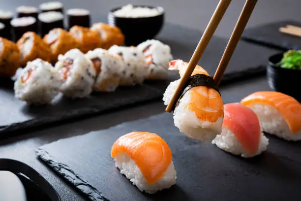 Shrimp sushi nigiri on a black plate with wooden chopsticks. Closeup of nigiri set with chopsticks and soy sauce with uramaki and hosomaki in background. Eating japanese food on black slate dishes at restaurant.