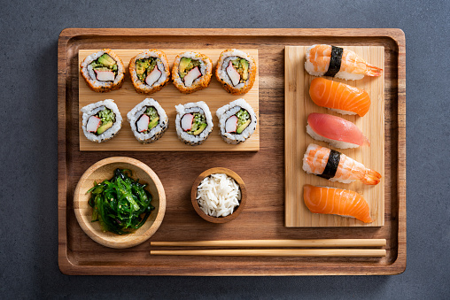 Top view of delicious variety sushi pieces setting on wooden tray. High angle view of japanese food with huramaki, maki, rolls, nigiri, seaweed and rice in a bowl. Flat image of fresh japanese sushi served on bamboo tray with chopstick on slate table.