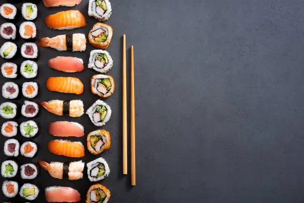 Top view of nigiri, hosomaki, uramaki and wooden chopsticks on black stone. High angle view of sushi rolls and maki on dark blackboard. Japanese food on dark background with copy space.