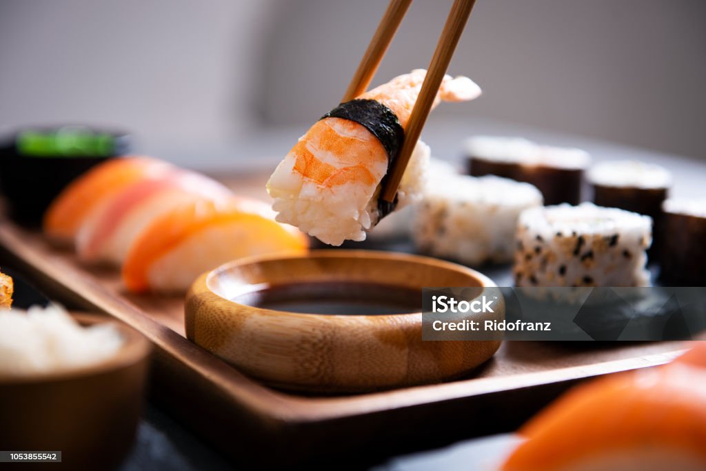 Chopstick with nigiri sushi piece Closeup hand holding bamboo chopsticks with nigiri shrimp while soaking it in soy sauce. Detail of sushi set on wooden tray at restaurant while hand dip nigiri in soy sauce. Japanese cuisine concept. Sushi Stock Photo