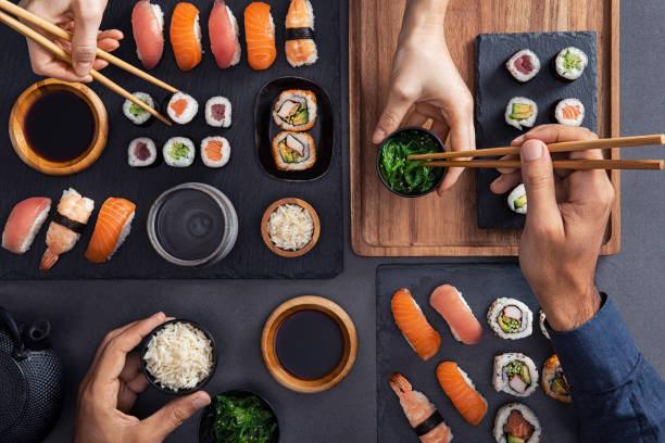 Sharing and eating sushi food Top view of couple hands eating sushi food at japanese restaurant. High angle view of woman hand serving seaweed in little bowl with sesame to man while holding hosomaki with chopsticks. Couple eating and sharing sushi roll, maki, nigiri, uramaki. japanese cuisine food rolled up japanese culture stock pictures, royalty-free photos & images