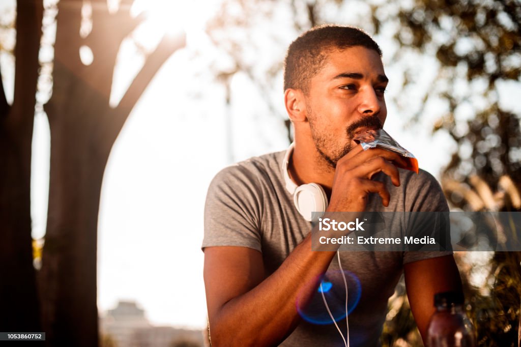 Young man eating candy bar African-American male with headphones eating chocolate candy bar while resting from outdoor exercise. Eating Stock Photo