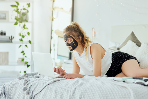 Cropped shot of an attractive young woman using her laptop while wearing a face mask in her bedroom at home