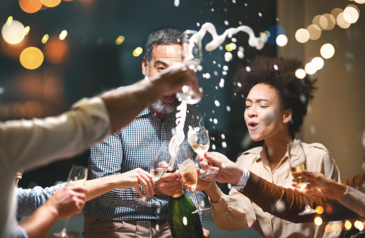 Closeup low angle view of group of mixed age adults toasting with wine at a house party. There are five men and three women. One of the men popping of the cork while the champagne is splashing all over.