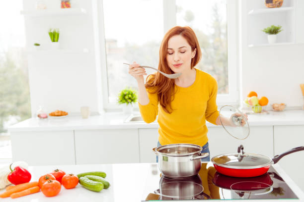 トマト、キュウリ、ピーマン、キャベツと、黄色のセーターの女性モダンな白い明るい居心地のよい、快適なインテリアのテーブル上にニンジンは、家族の朝食のおいしい料理を作る - chef cooking food pan ストックフォトと画像