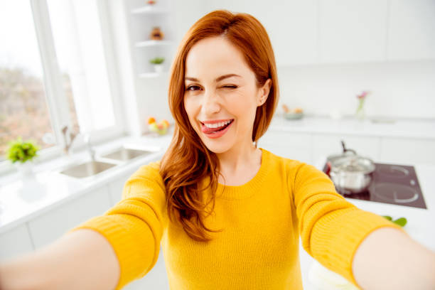 istock Elegant, graceful, charming, cheerful, fancy woman in yellow pullover take selfie on front camera of smartphone make tongue out wink eye in modern white bright interior 1053867550