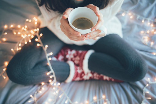 Woman sitting at home legs crossed on bed, holding a cup of coffee, christmas decoration.
