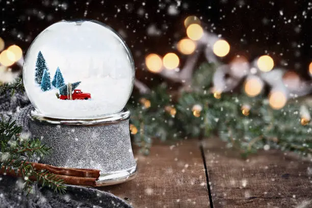 Rustic image of a snow globe with old pick up tuck hauling a Christmas tree surrounded by pine branches, cinnamon sticks and a warm gray scarf with gently falling snow flakes.