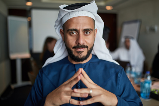 Portrait of confident Arab entrepreneur at conference table with colleagues in background. Confident Emirati male professional is sitting with hands clasped in board room. He is wearing kaffiyeh at workplace.