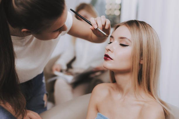Makeup Artist with Brush in Hand and Blond Model. Blond Girl in Beauty Salon. Makeup in Beauty Salon. Makeup Artist with Brush. Woman and Makeup Artist. Beautiful Woman. Blond Girl with Shining Skin. Blond Girl. Model and Makeup Artist. Red Lips. makeup artist stock pictures, royalty-free photos & images