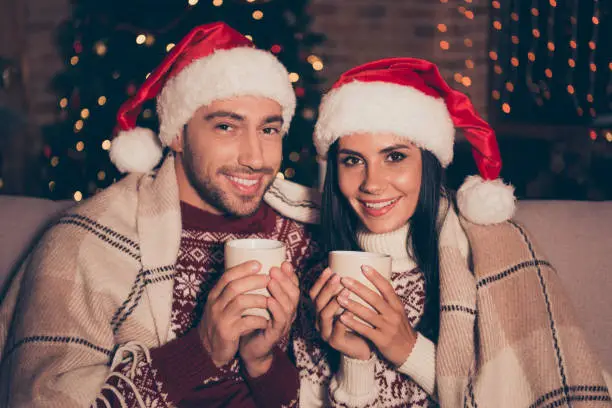 Two wonderful, adorable, good-looking, people in soft ornament sweater, cover blanket hold hot aroma mulled-wine beverage hands sit on cozy couch in living room with sparkles garland decorations