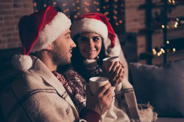 Two good-looking, beautiful, dream, sweet, charming, attractive, dreamy, cute people in ornament sweater sit on comfort, cozy couch in living room with mulled-wine drink in hands look to each other