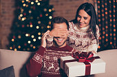 Hide and seek concept. Good-looking, attractive brunette lady in ornament sweater hold big package with bow, close eyes her man, who sit in cozy living room couch with lights garland decorations