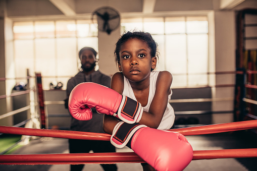 Confident looking boxing kid standing inside a boxing ring with her coach in the background. Girl wearing boxing gloves standing while resting her hands on the boxing ring.