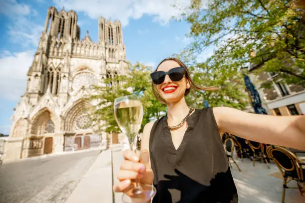 Young woman making selfie portrait with champagne standing in front of the cathedral in Reims city, capital of Champagne wine region, France