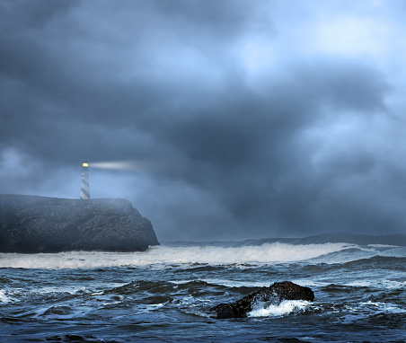 A lighthouse sits on top of  a rocky cliff that overlooks a rough ocean in foggy and inclement weather.  The cloudy and foggy sky provide ample room for copy and text.