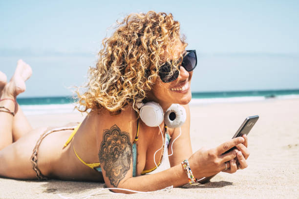 sexy beautiful curly hair caucasian middle age lady in bikini laying at the beach with the blue ocean in background. vacation leisure activity listening music with headphone and phone. cheerful cute lady in vacation enjoying summer - headphones women tattoo music imagens e fotografias de stock
