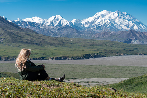 Beautiful blonde woman sits on a hill looking out at Mount Denali (McKinley) in Denali National Park. Concept for thinking, peaceful, nature, exploring