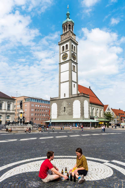 Augsburg, Germany - August 18, 2018: City of Augsburg Town Hall. Two young people drinking drinks sitting on the floor in the city square of Augsburg. This city is located in Bavaria, Germany. The building designed and erected between 1615 and 1624 by Eliot Holl, master builder of the city. The site is protected by the Hague Convention for the Protection of Cultural Property in the Event of Armed Conflict because of its cultural and historic importance.