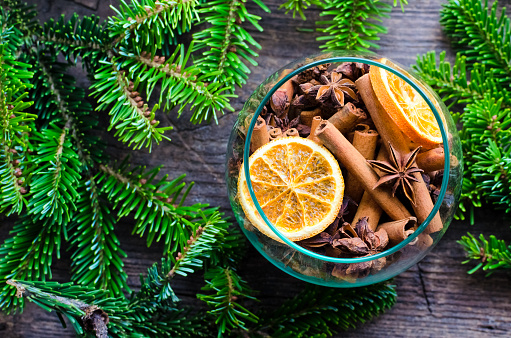 Christmas fragrance concept. Christmas spices still life with cinnamon sticks, star anise and dried orange slices in glass bowl on a wooden background with fir tree. Top view.