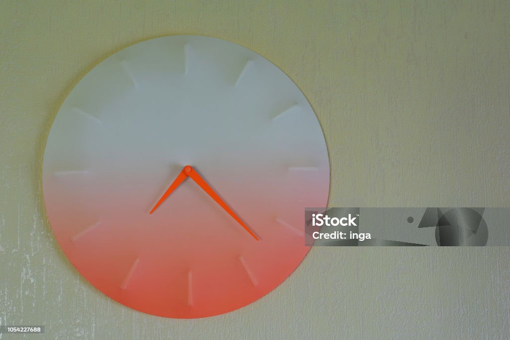 Round orange clock hanging on light yellow wall Round orange clock hanging on light yellow wall background with copy space for text. Alarm Stock Photo