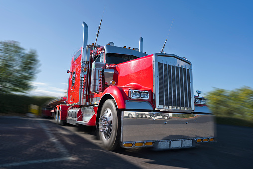 Front view of red classic bonnet American big rig semi truck tractor with awesome stylish chrome accessories standing on truck stop with flat bed semi trailer caring covered commercial cargo