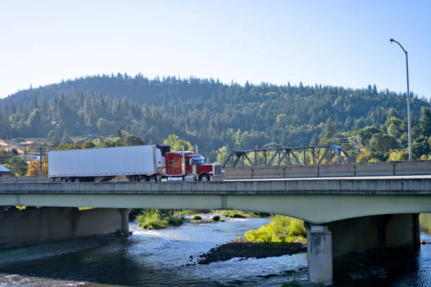 istock Red classic big rig semi truck with refrigerated semi trailer transporting cargo on interstate highway with bridge across the river 1054227948