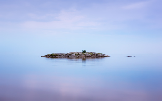 A small stony island on a calm water surface of a lake reflecting misty sky in a summer northern night. Amazing seascape background with copy space. White Nights season, Karelia, Russia.