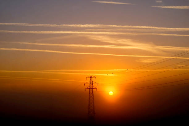 electricity pylon silhoutted by the low yellow sun that is rising through the centre of the pylon there is mist on the ground the morning sky is covered with sixteen white vapor trails contrails from airoplanes running horizontally accross through the mist in the distance is another electricity pylon with power cables