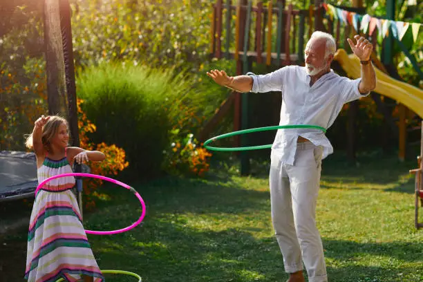 Multi generation family hula hooping in backyard. Senior man and his granddaughter having fun outdoors.