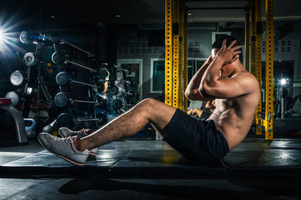 chico musculoso haciendo sit ups en el gimnasio con los demás en segundo plano. atleta joven haciendo ejercicios de estómago en el gimnasio moderno. ajuste guapo haciendo abdominales en el gimnasio. - sit ups fotografías e imágenes de stock