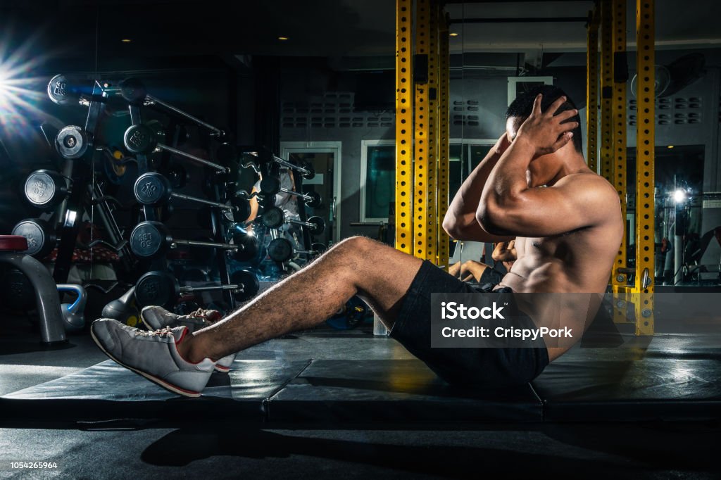 Muscular guy doing sit ups at gym with other people in background. Young athlete doing stomach workout in modern gym. Handsome fit man doing crunches at gym. Gym Stock Photo