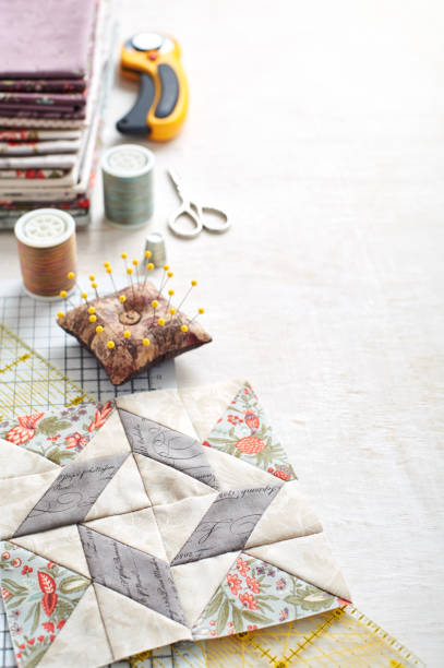 Patchwork block on craft mat, stack of fabric, sewing accessories on white wooden surface