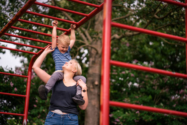 Woman carrying son on shoulders below monkey bars Low angle view of mother carrying boy on shoulders at public park. Mid adult woman is with stoma bag assisting son on monkey bars. They are playing outdoors against trees. jungle gym stock pictures, royalty-free photos & images