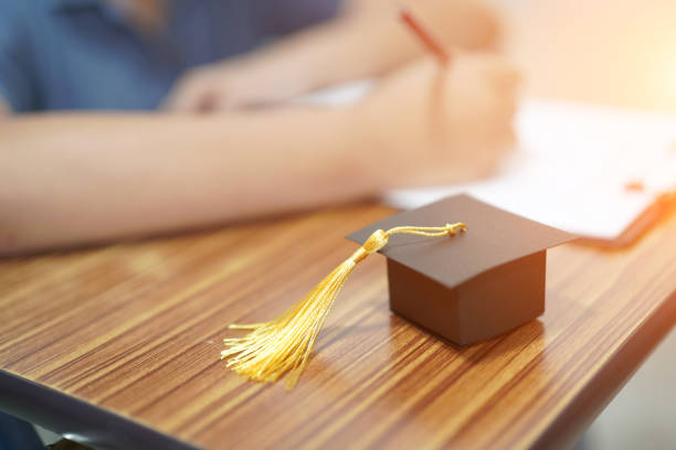 istock Asian children young kid write answer in a quiz examination test study learn with graduation gap hat on wooden table in school : bright genius education concept. 1054359062