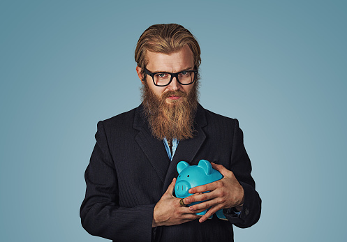Young greedy stingy Bearded hipster businessman man holding piggy bank Isolated on blue studio wall Background. Negative face expression human emotion body language reaction attitude. Horizontal
