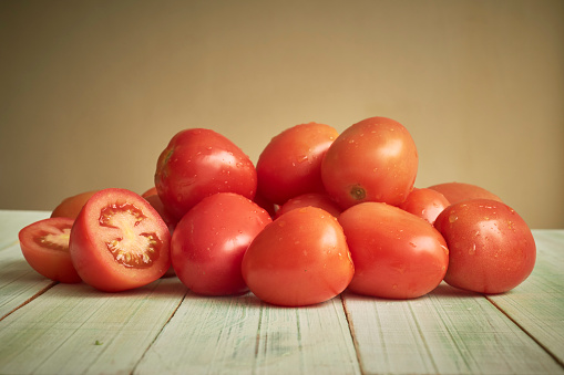 Heap of ripe raw tomatoes on rustic wooden table. One tomato is cut. Studio shot with natural day lighting. Front view. Close-up. Indoors.