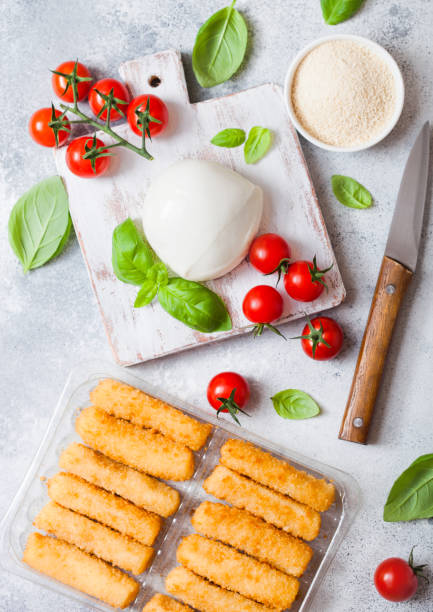 istock Fresh Mozzarella cheese on vintage chopping board with tomatoes and basil leaf and tray with cheese sticks on stone kitchen table background. 1054493832