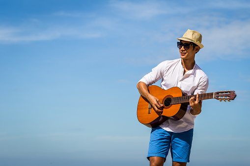Happy young Asian man playing classical guitar and standing over blue sky in background, copy space