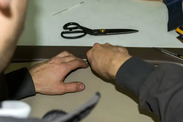 A male worker does manual work on skinning a genuine leather of a vehicle's interior with a beige-colored door to carry out a design solution in a car repair shop at the workplace with scissors