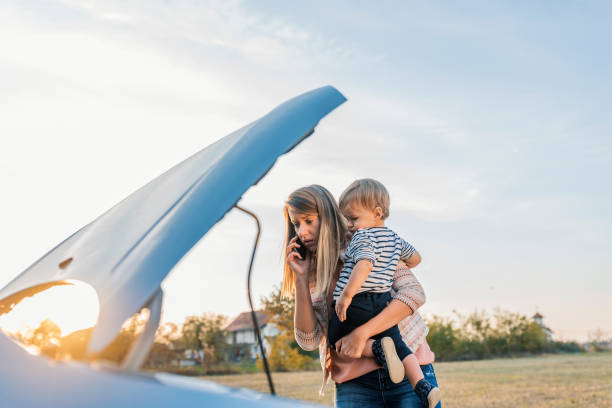 When can you get here? Portrait of young Caucasian woman standing by broken down car and waiting for assistance while holding her baby boy during sunset and talking on cell phone. Photo of female waits for assistance near her car broken down on the road side, using mobile phone. Frustrated young brown hair woman looking at broken down car engine on street, standing next to broken car and talking on the smartphone. Woman does not understand why her car engine failed. Desperate mother holding her angry baby. Young woman with broken car calling for help. broken spring stock pictures, royalty-free photos & images