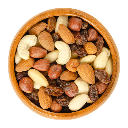 Nuts and raisins in wooden bowl. Snack mix of dried almonds, hazelnuts, cashews and raisins.\nTrail mix. Edible, raw, organic and vegan. Isolated macro food photo closeup from above on white background